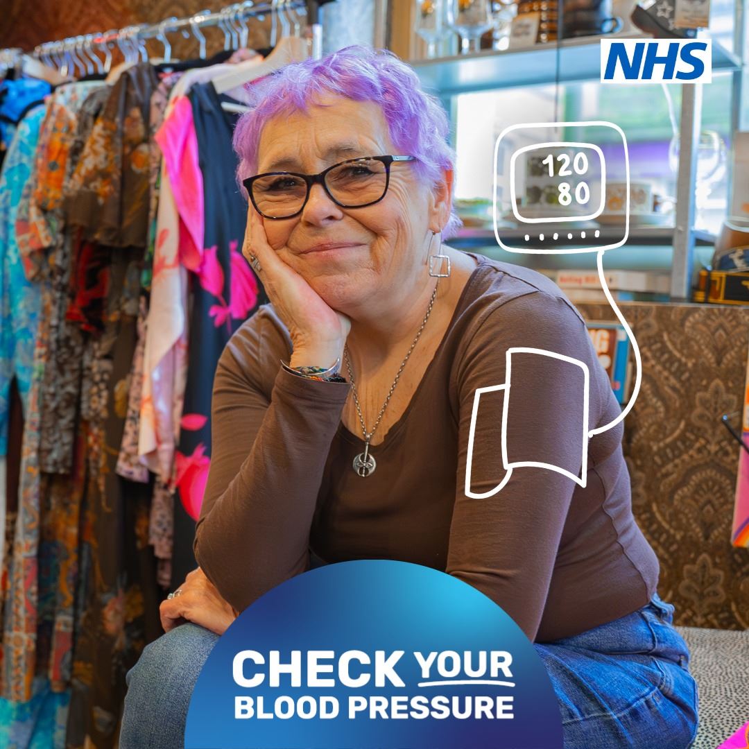 a middle aged woman with short hair dyed purple.  She is sat in the dressing room in a clothes store.  A carton image of a blood pressure monitor is drawn over her left arm.  The NHS logo appears at the top right and the words Check Your Blood Pressure appear at the bottom