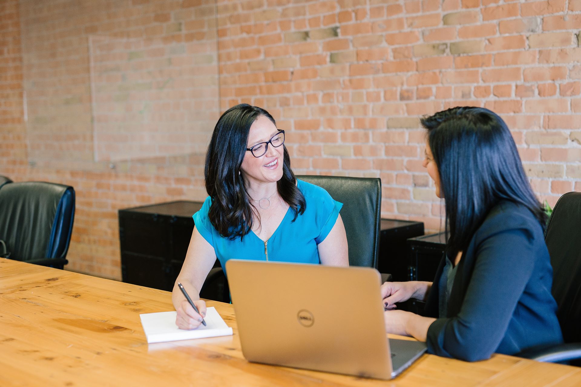 two woman sat at a table with a laptop, and writing on pad