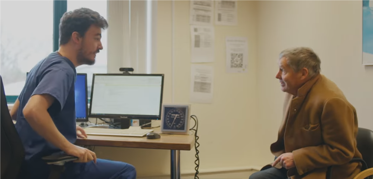 a healthcare assistant speaking with an older man in a GP clinical room